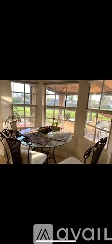 A dining table with chairs and a glass top is set in a room with large windows.