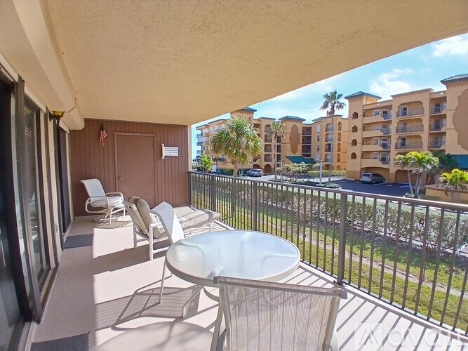 A balcony with a table and chairs overlooks a parking lot and apartment buildings.