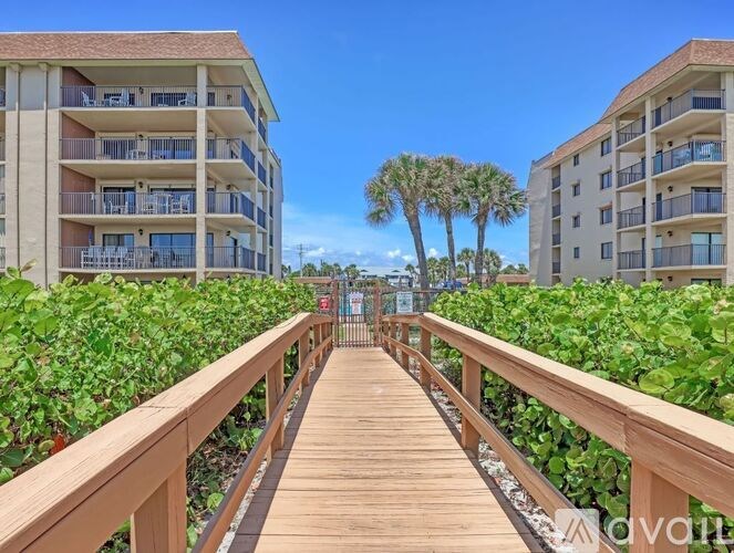 A wooden walkway leads between two apartment buildings with lush greenery on either side.
