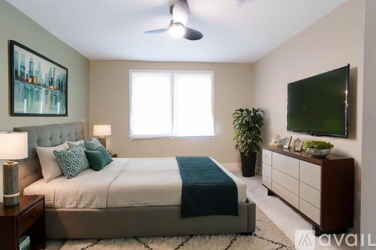 A bedroom with a bed, a ceiling fan, a plant, and a painting on the wall.