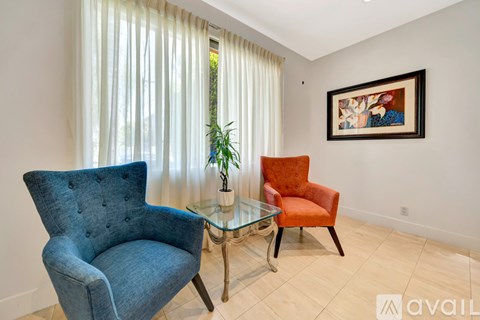 A blue chair and an orange chair are placed in front of a glass table and a framed picture on the wall.