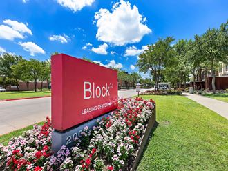 a red block sign in front of a garden of flowers