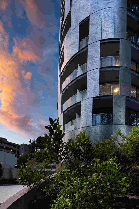 A tall building with balconies and windows is surrounded by greenery.
