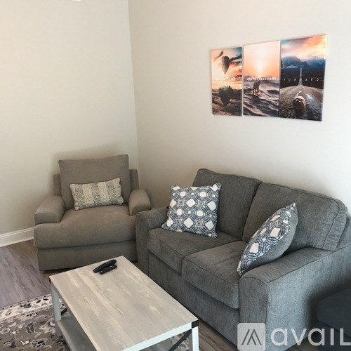 A living room with a grey couch, a grey armchair, a coffee table, and a wall with a picture of a landscape.