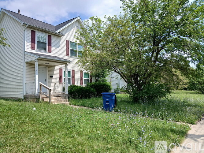 A white house with red shutters and a blue trash can in front of it.