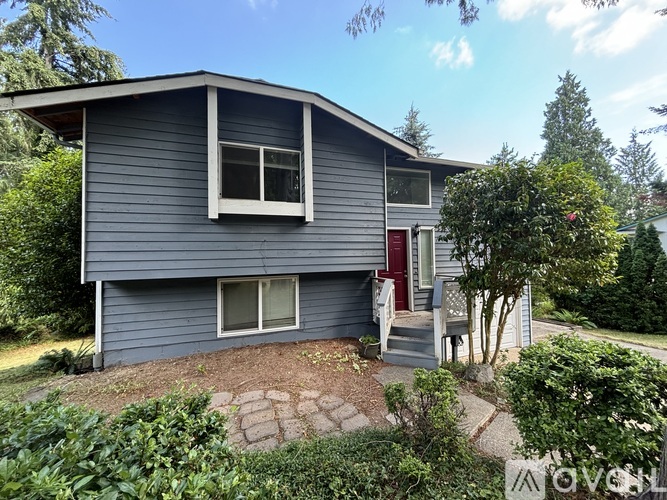 A house with a grey exterior and a red door is surrounded by greenery.