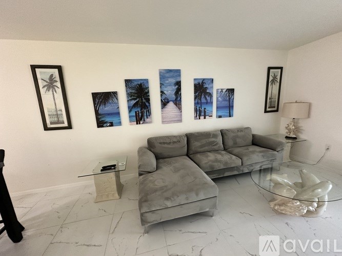 A living room with a grey couch, a glass coffee table, and a wall with four pictures of palm trees.