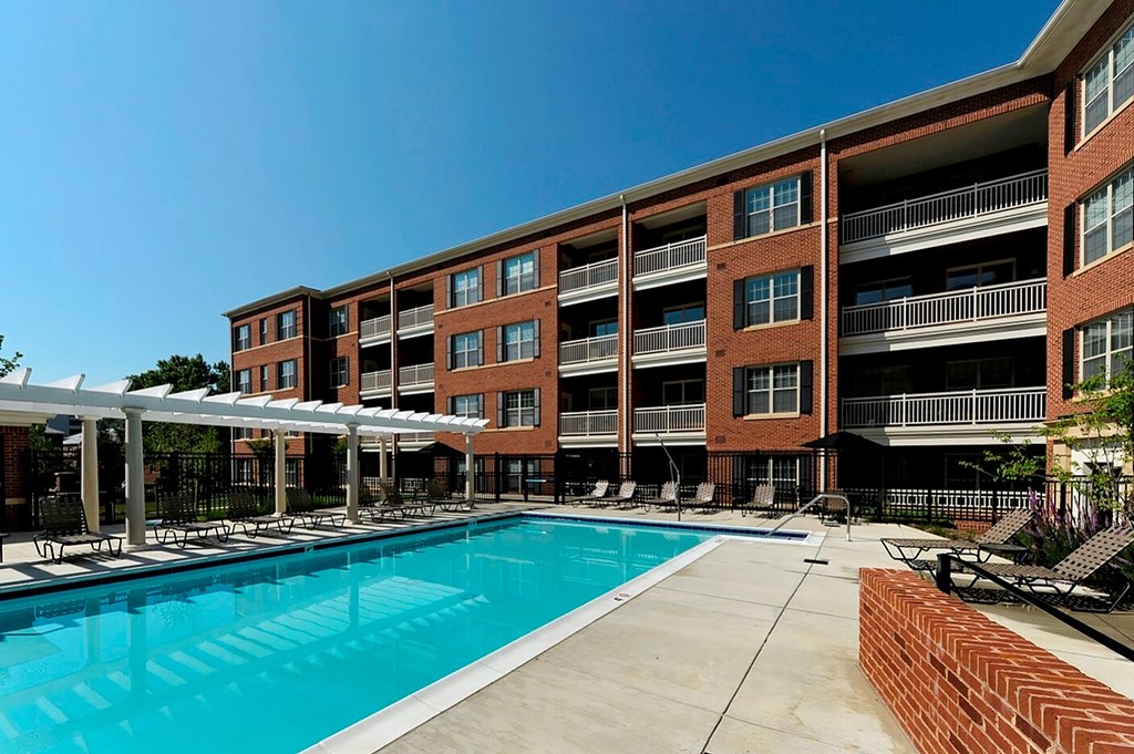 A large swimming pool is surrounded by a covered patio area and a brick building.