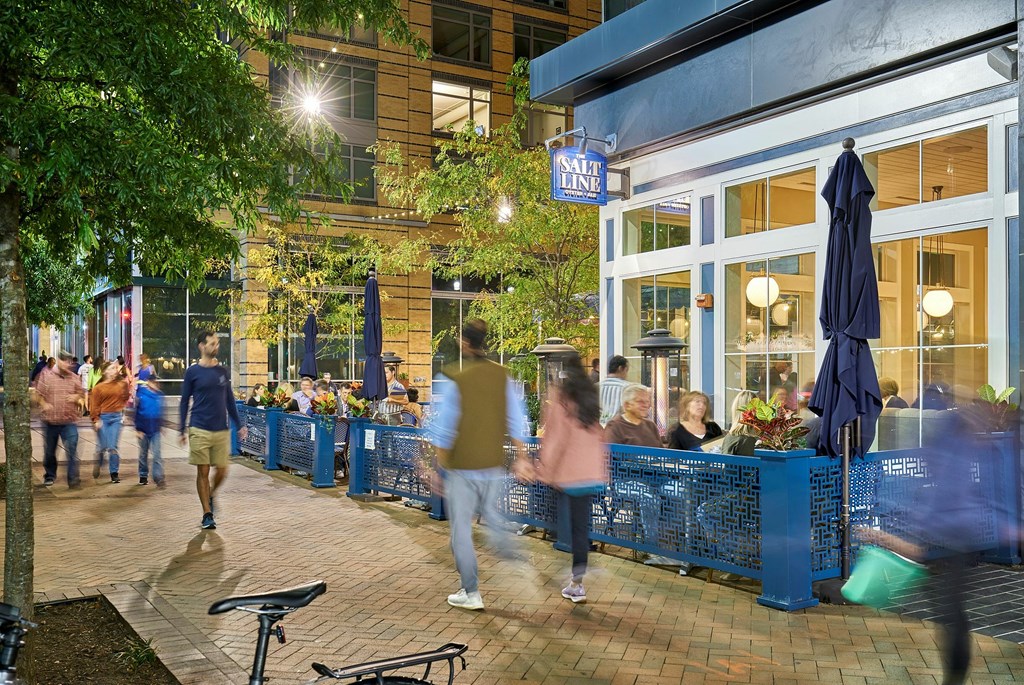 A busy outdoor patio scene with people walking and sitting at tables.