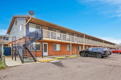 A black car is parked in front of a two-story apartment building with a balcony and stairs.