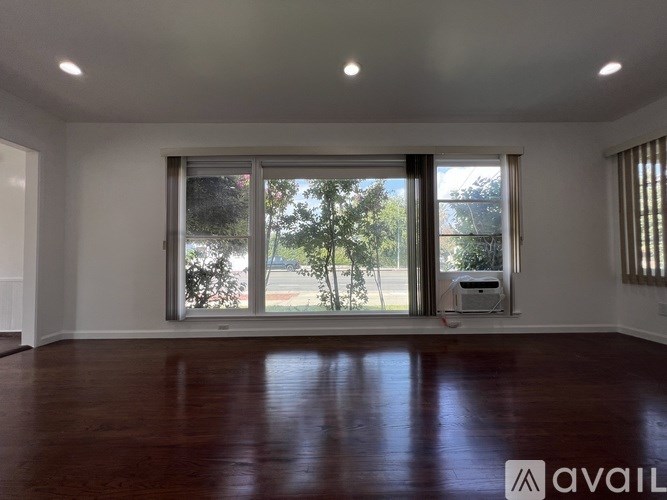 A room with wooden floors and a large window with a view of trees and a printer on the floor.