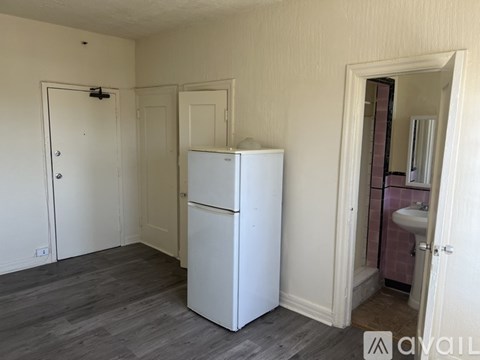 A white refrigerator is in the corner of a room with a sink and mirror in the background.