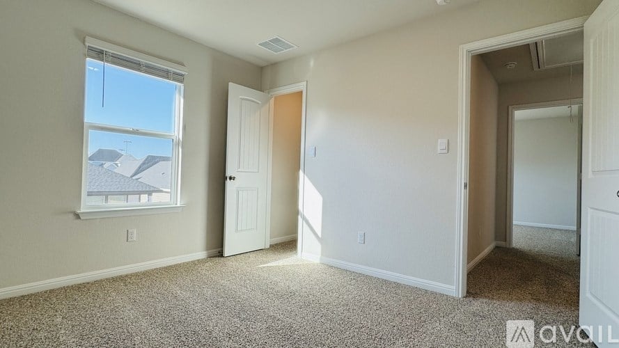 A room with a carpeted floor, a window with a view of houses, and a door leading to another room.