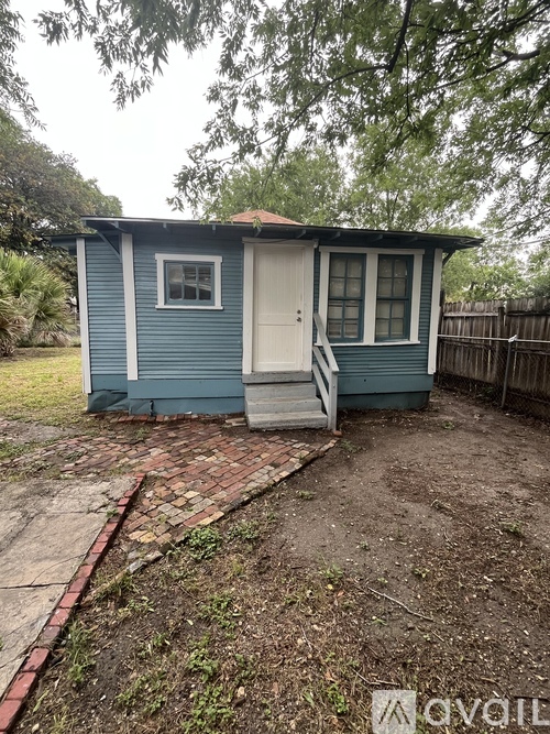 A small blue house with a white door and windows is surrounded by a fence and trees.