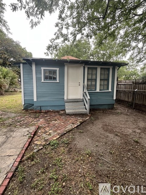 A small blue house with a white door and windows is surrounded by a fence and trees.