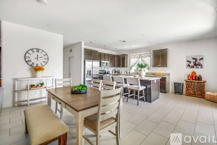A dining room with a wooden table and chairs, a clock on the wall, and a kitchen area in the background.