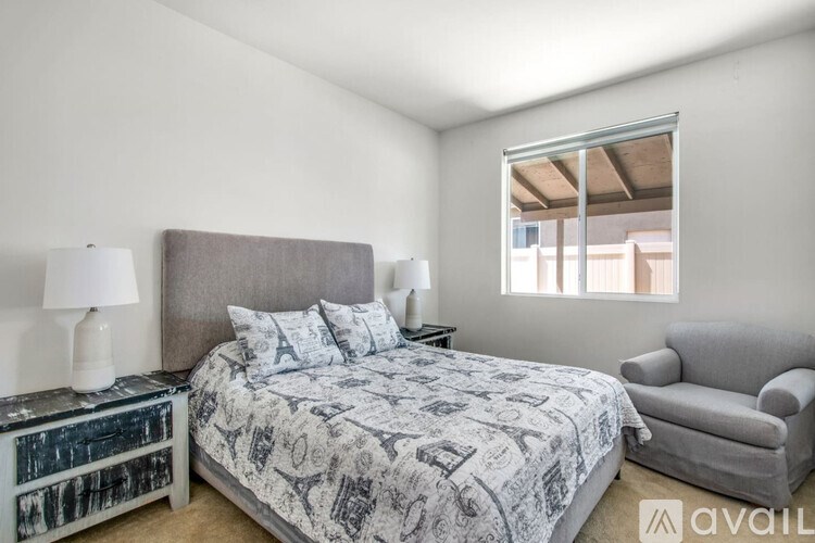 A bedroom with a patterned bedspread, a grey headboard, a grey armchair, and a nightstand with a lamp.