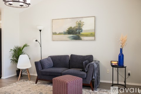 A living room with a grey couch, a red ottoman, a white floor lamp, and a painting on the wall.