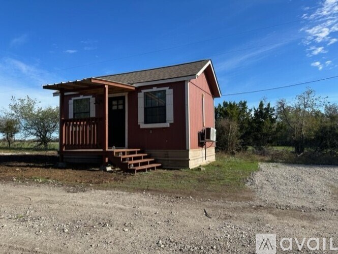 A small red house with a porch is situated in a gravel area with trees in the background.
