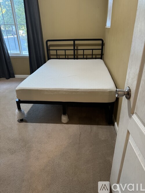 A bed with a white mattress and a black metal frame is in a room with a carpeted floor and a window with curtains.