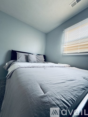 A bed with a white comforter and two pillows sits in a room with a window covered by blinds.