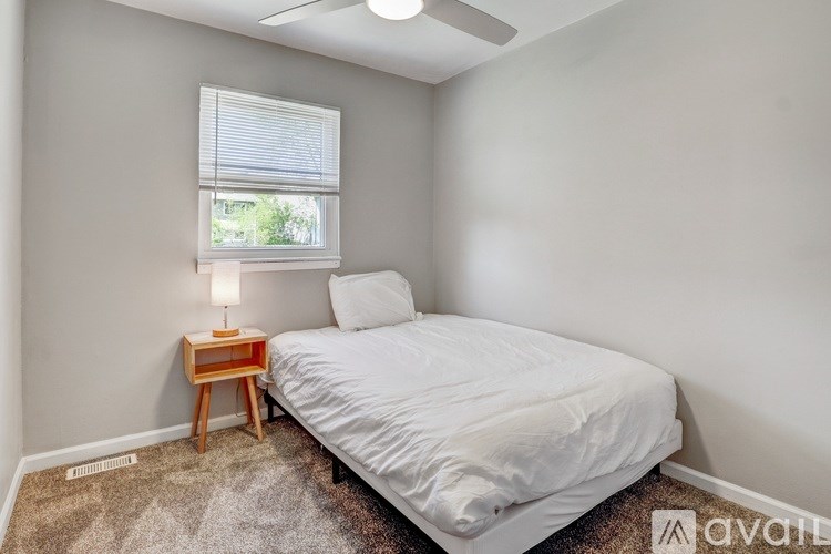 A bedroom with a bed, a window with blinds, a small table with a lamp, and a carpet.