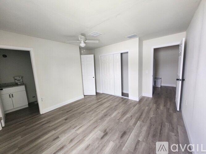 A room with a wooden floor and white walls, with a ceiling fan and a door on the right.