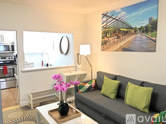 A living room with a grey couch, a vase with pink flowers on the coffee table, and a painting of a street scene on the wall.