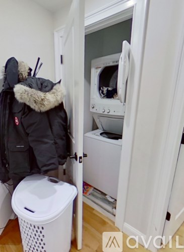 A white washing machine is in a small room with a white trash can and a furry jacket hanging on the wall.