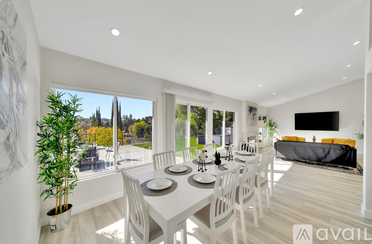 A dining room with a white table and chairs, a television, and a view of the outdoors through large windows.