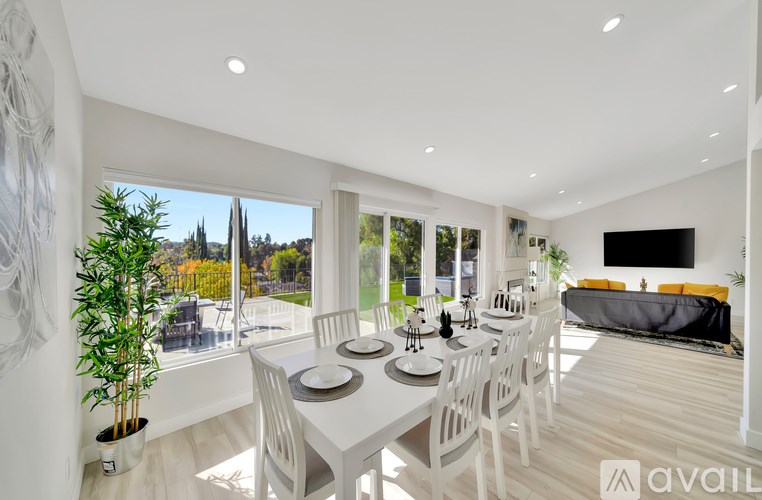 A dining room with a white table and chairs, a television, and a view of the outdoors through large windows.