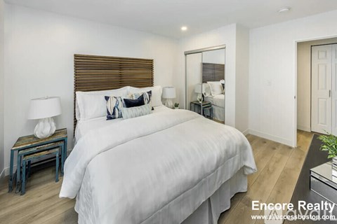 A bedroom with a large bed, a nightstand with a lamp, and a mirror on the wall.