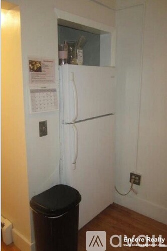 A small white refrigerator in a room with a calendar and a black trash can.