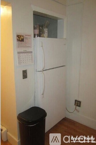 A small white refrigerator in a room with a calendar and a black trash can.