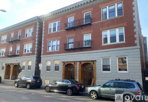 A row of red brick buildings with balconies and cars parked in front.