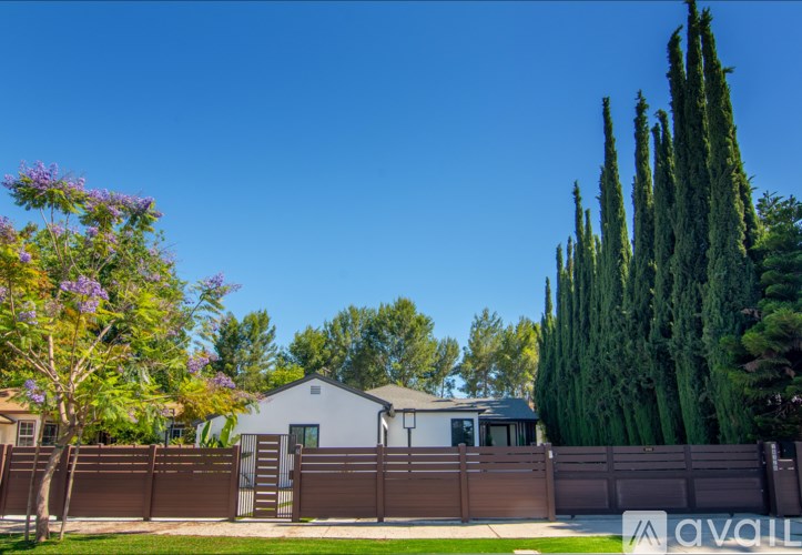 A white house with a brown fence and a tall green tree in front.