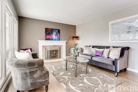 A living room with a grey couch, a grey armchair, a glass coffee table, and a fireplace with a painting above it.