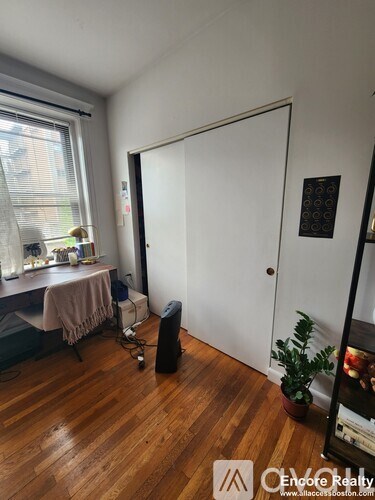 A room with a wooden floor, a desk with a computer, a chair, a window with blinds, a white door, a black speaker, and a plant.