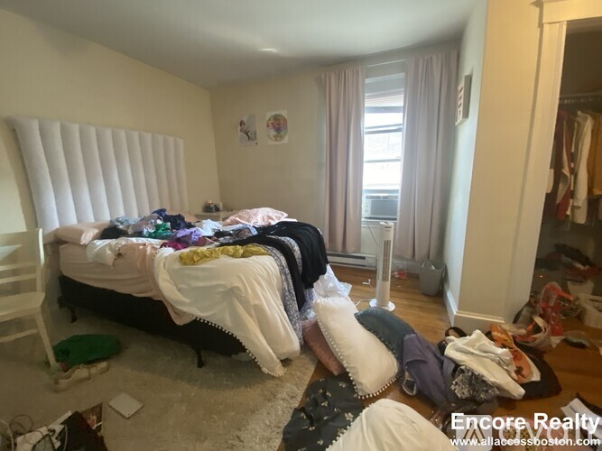 A messy bedroom with a bed covered in clothes and a fan on the floor.