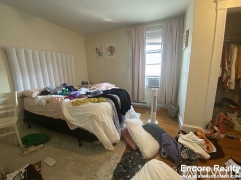 A messy bedroom with a bed covered in clothes and a fan on the floor.