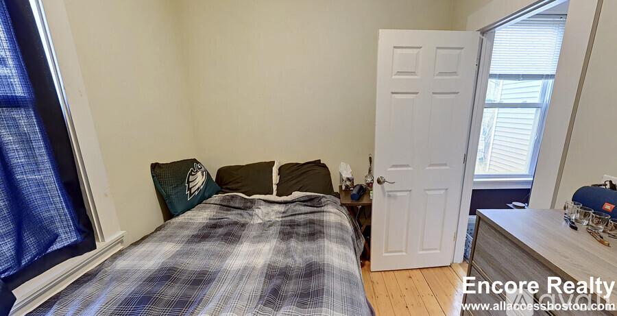 A bedroom with a bed, a window with curtains, a dresser with a lamp, and a door.