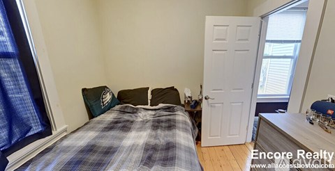 A bedroom with a bed, a window with curtains, a dresser with a lamp, and a door.