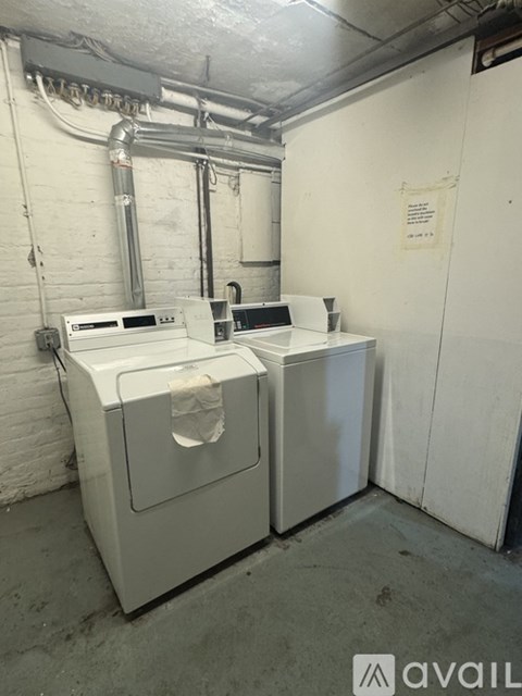 A white industrial washing machine is in a concrete room with pipes and a sign on the wall.