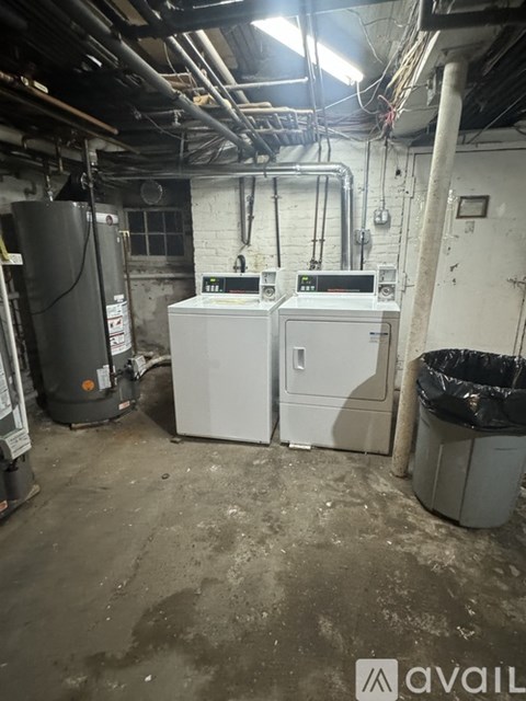 A dirty basement with a refrigerator, two white boxes, and a trash can.