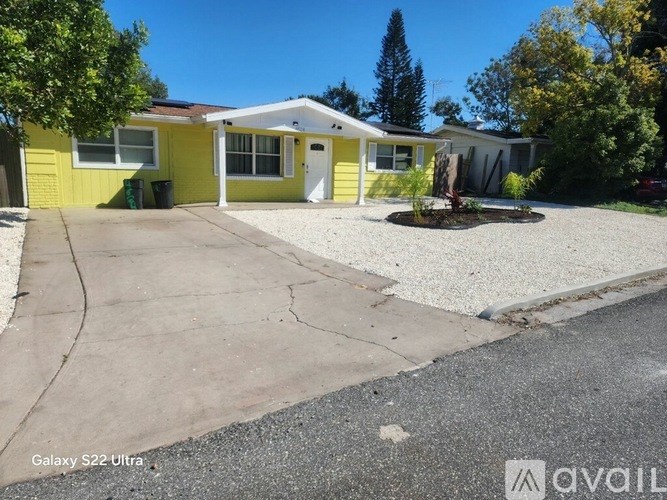 A yellow house with a white door and windows is surrounded by trees and has a gravel driveway.