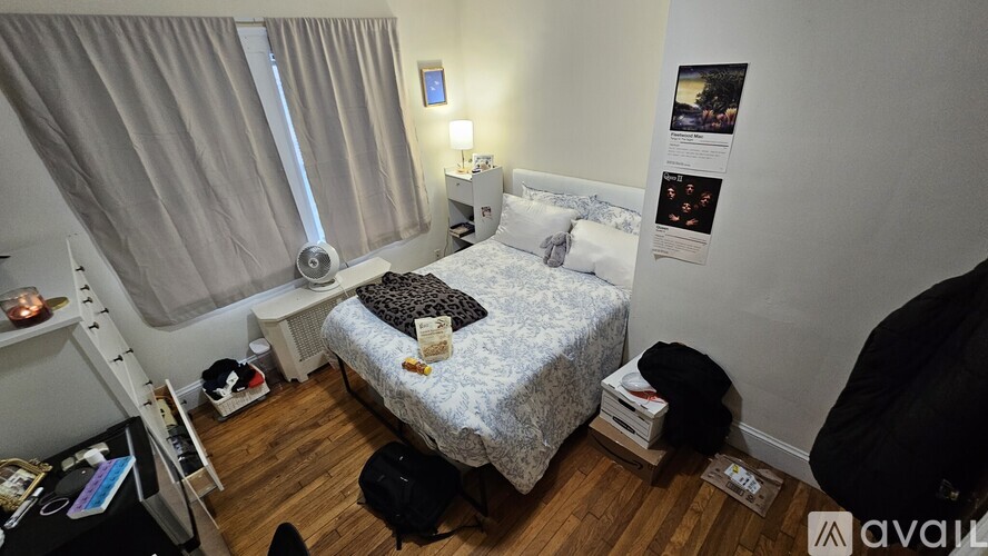 A small bedroom with a bed, a window with curtains, a nightstand with a lamp, and a wall with pictures and posters.