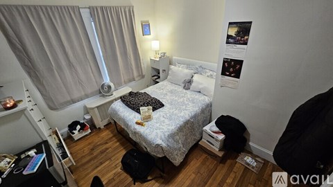 A small bedroom with a bed, a window with curtains, a nightstand with a lamp, and a wall with pictures and posters.