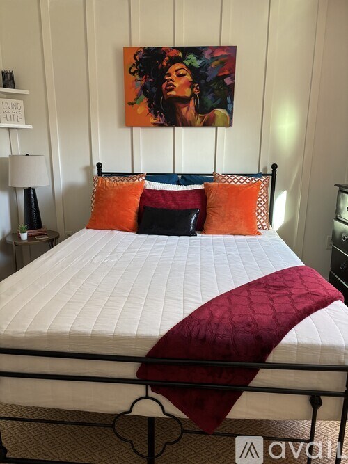 A bed with a white comforter and a red blanket is in the foreground with a painting on the wall behind it.