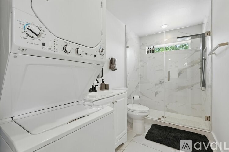 A white washing machine is in a small bathroom with a toilet and a glass shower stall.