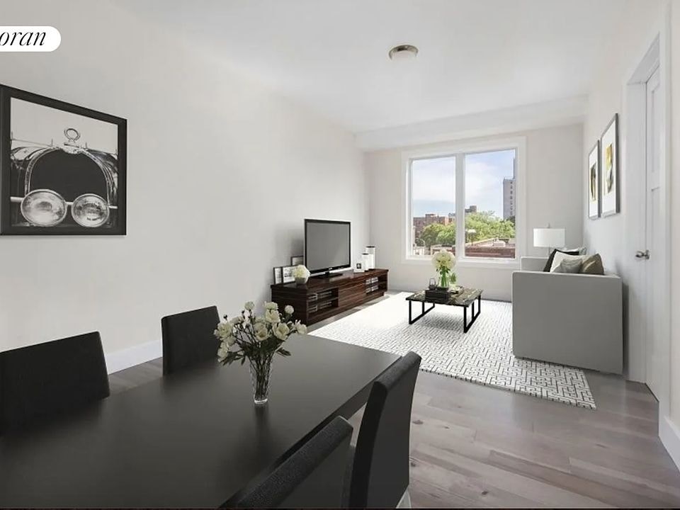 A black dining table with four chairs and a vase of flowers is in the foreground of a well-lit room with a large window and a grey sofa in the background.